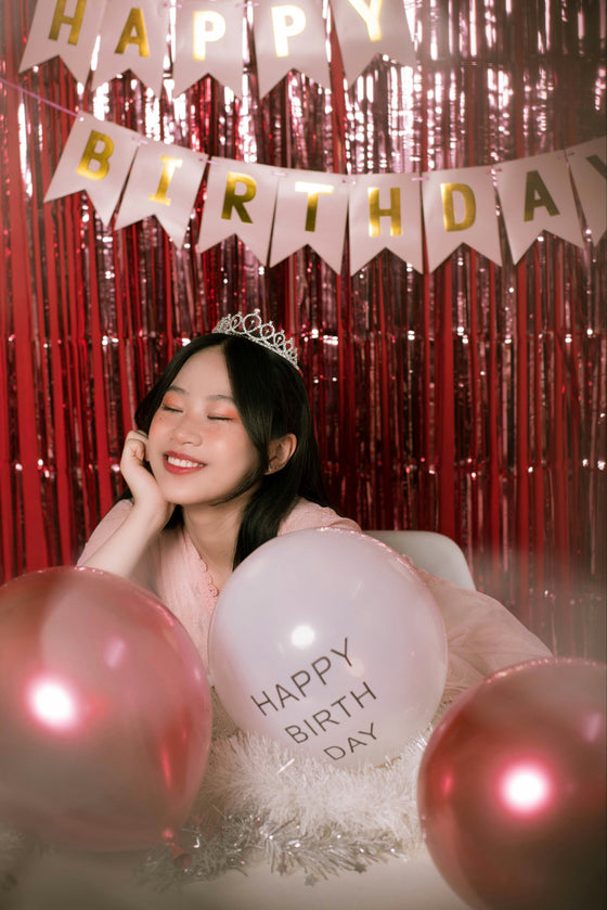 A woman wearing a tiara sits in a celebratory birthday setup with pink metallic tinsel curtains, a 'HAPPY BIRTHDAY' banner, and pink balloons, including one that reads 'HAPPY BIRTH DAY'.